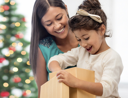 A woman smiles as a girl opens a gift in front of a Christmas tree